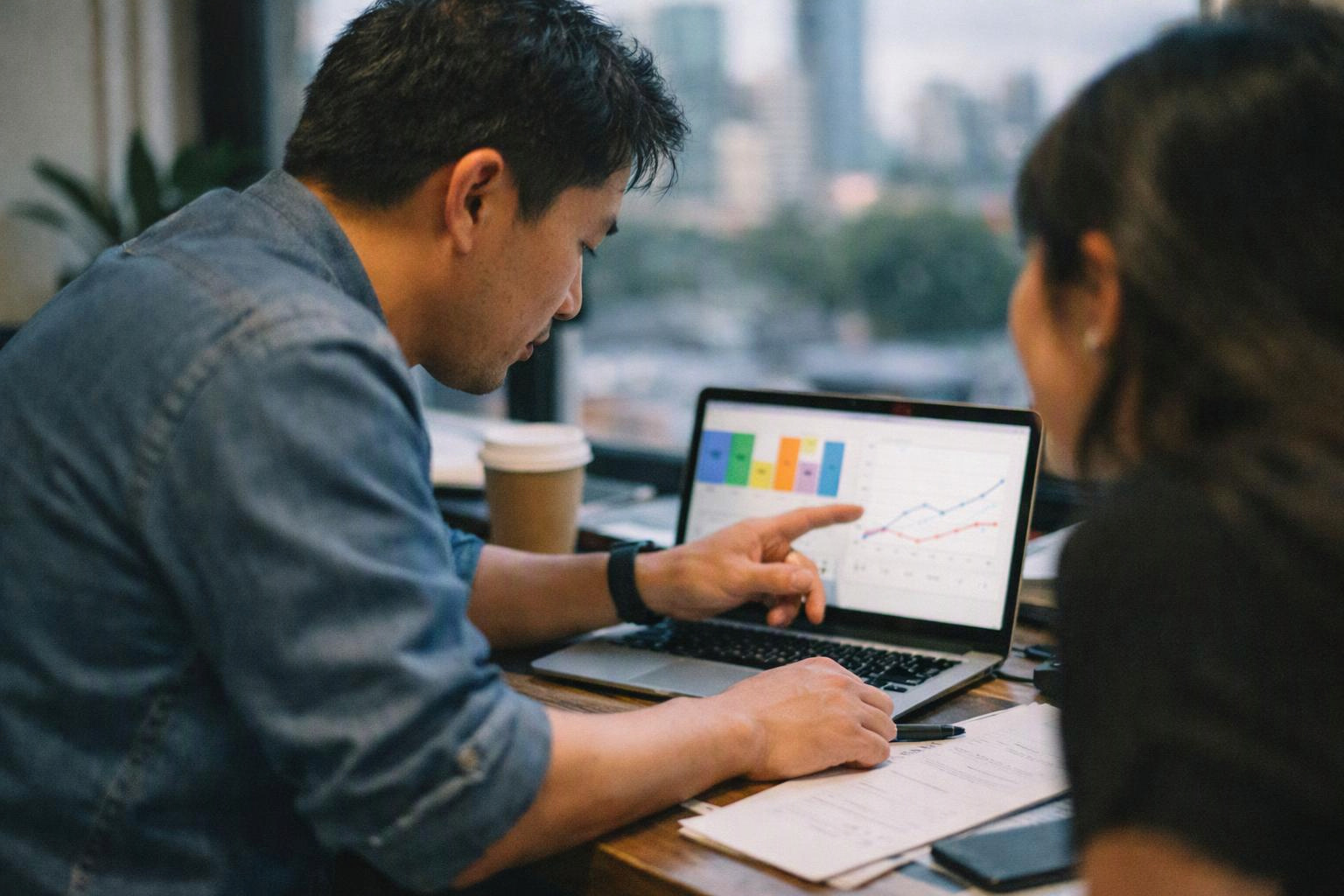 Two coworkers sit at a table, studying charts on a laptop beside coffee and papers