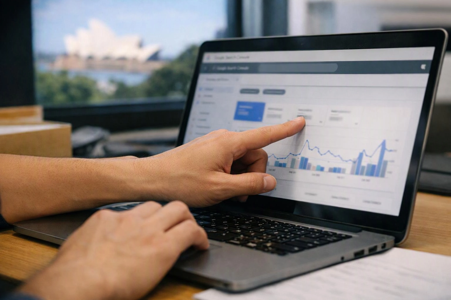 A person points at a laptop analytics chart on a desk by a window