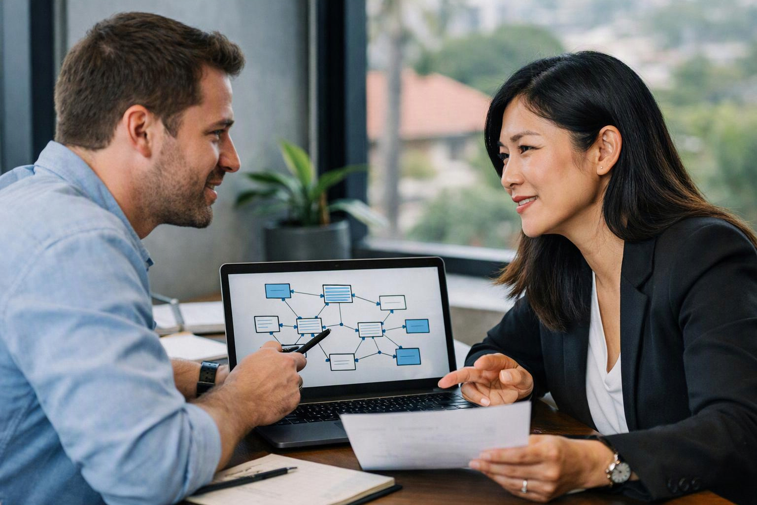 Two coworkers discuss a laptop flowchart at a desk near large windows