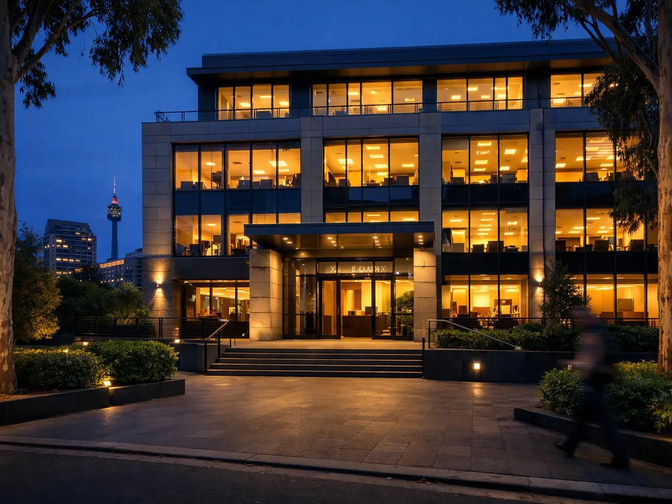 A lit office building stands at dusk, with glowing windows, steps, trees, and a blurred person walking past