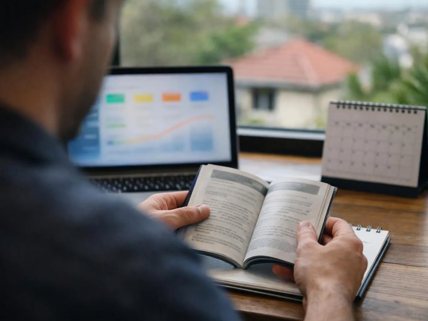 A person reads an open book at a desk beside a laptop, calendar, and notebook near a window