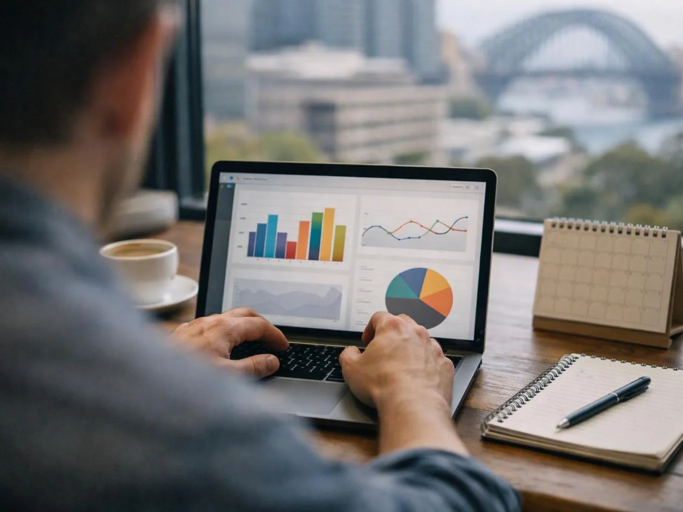 A person works on a laptop showing colorful charts at a desk by a window with a city view