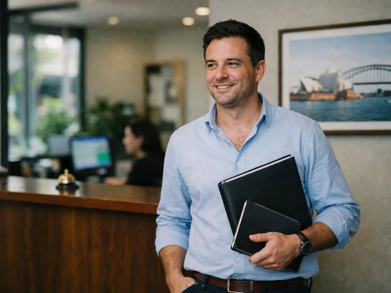 Smiling man in a light blue shirt holds folders beside a reception desk in an office lobby