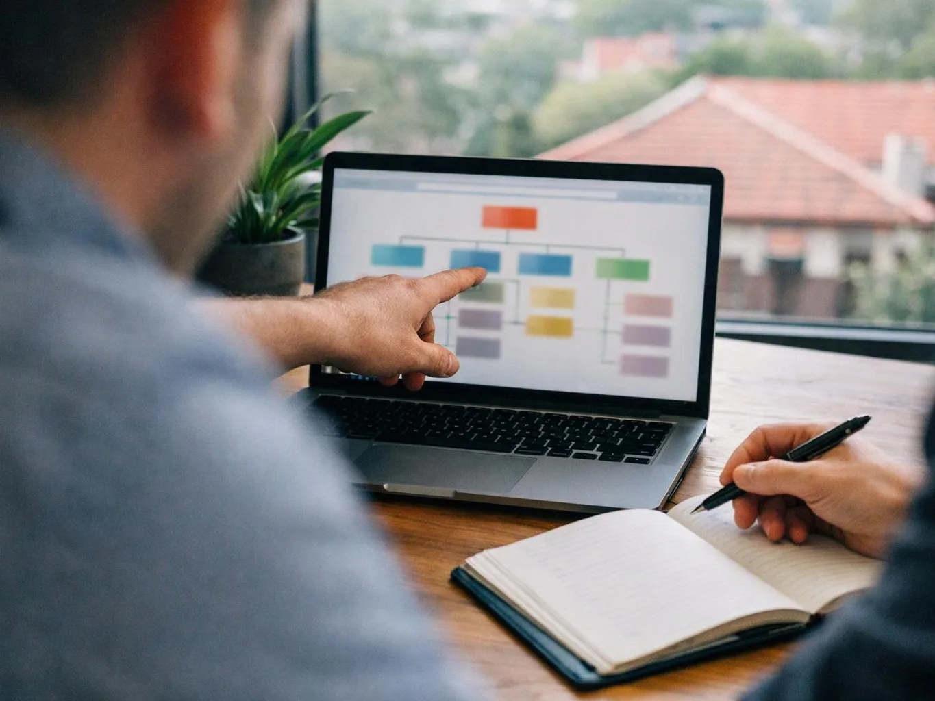 Two people review a colour flowchart on a laptop while one points and the other writes in a notebook