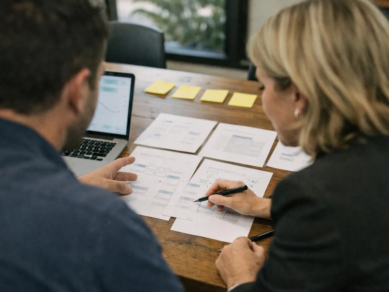Two people review printed charts and wireframe pages beside a laptop at a wooden table